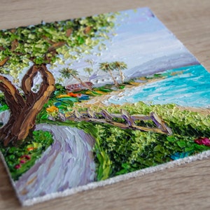 K&ouml;nnte beinhalten: Ein Gem&auml;lde, das eine Strandszene mit einem Weg zum Ufer zeigt. Der Weg ist von gr&uuml;nen B&uuml;schen und einem Holzzaun ges&auml;umt. Ein gro&szlig;er Baum mit gr&uuml;nen Bl&auml;ttern befindet sich auf der linken Seite des Gem&auml;ldes. Der Strand ist sandig mit blauem Wasser und wei&szlig;en Wellen. Der Himmel ist hellblau mit wei&szlig;en Wolken.