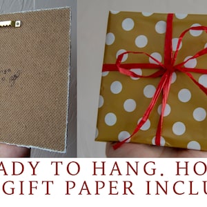 K&ouml;nnte beinhalten: Ein kleines, gerahmtes Kunstwerk mit einem Haken auf der R&uuml;ckseite, bereit zum Aufh&auml;ngen. Es ist in goldenes Geschenkpapier mit wei&szlig;en Punkten und einem roten Band verpackt.