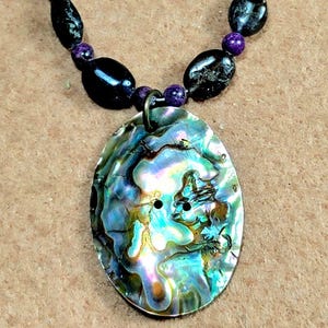 May include: Abalone shell pendant necklace with a pearlescent, iridescent surface. The oval pendant is suspended from a beaded chain of black and purple stones. The necklace has a bronze-colored clasp.