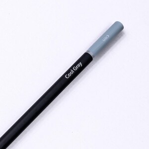 May include: A sharpened pencil with a black barrel featuring the white text Cool Gray. The end of the pencil is capped with a light gray color indicator. The pencil has a wooden casing visible at the sharpened tip with a dark gray lead core.