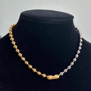 May include: A two-tone necklace with a ball chain design. One half of the necklace is gold-colored, and the other half is silver-colored. The necklace is displayed on a black mannequin.