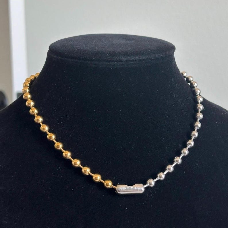 May include: A two-tone necklace with a ball chain design. The necklace is gold on one side and silver on the other, connected by a silver clasp with small, clear stones. The necklace is displayed on a black mannequin.