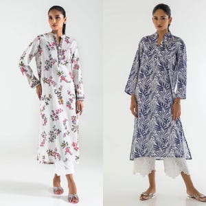 May include: Two women wearing long-sleeved tunics with floral prints. The tunic on the left is white with pink and blue flowers. The tunic on the right is blue with white leaf patterns. Both women are wearing white pants.