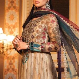 Pode incluir: Uma mulher vestindo um vestido longo e fluido de cor dourada e creme com bordados e enfeites intrincados. Ela est&aacute; usando um dupatta combinando com uma borda colorida. O vestido tem um corpete justo e uma saia fluida.