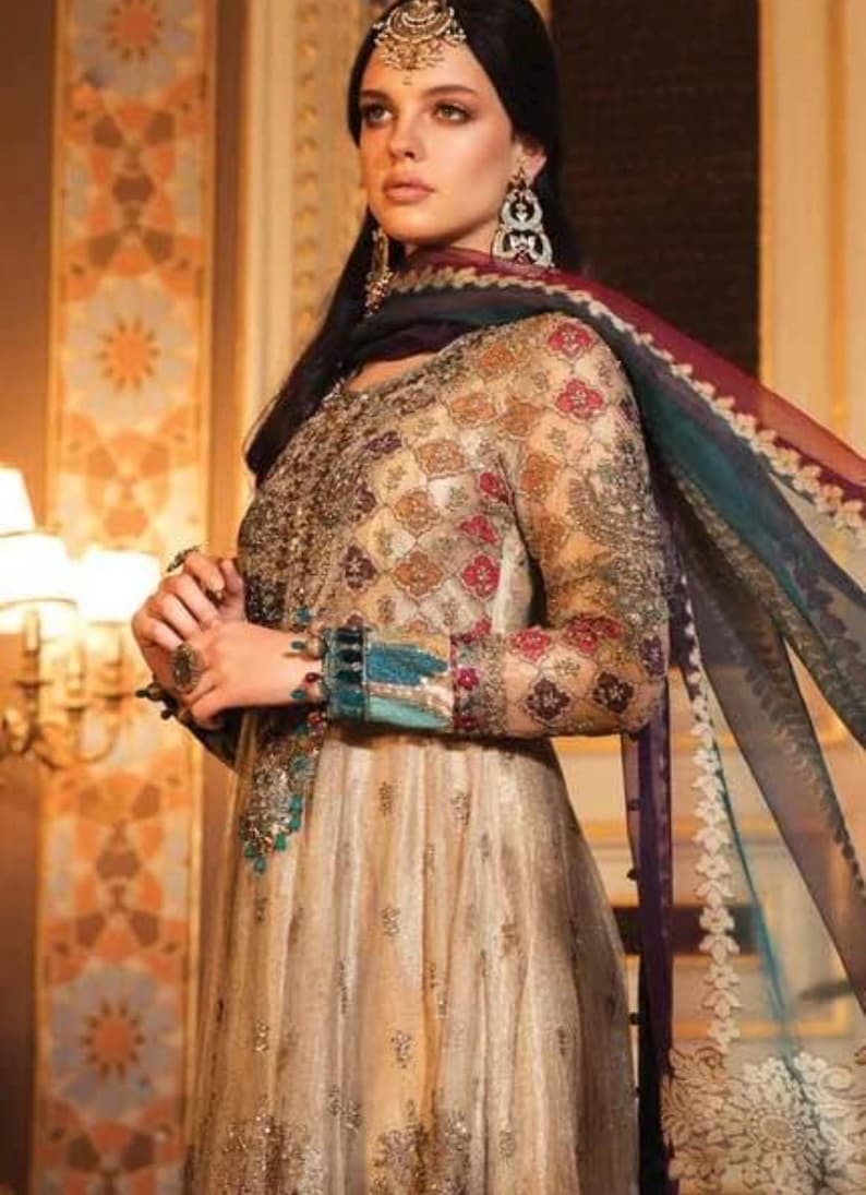 Pode incluir: Uma mulher vestindo um vestido longo e fluido de cor dourada e creme com bordados e enfeites intrincados. Ela est&aacute; usando um dupatta combinando com uma borda colorida. O vestido tem um corpete justo e uma saia fluida.