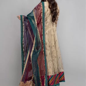 Pode incluir: Um vestido longo e fluido de cor bege com bordados dourados e uma bainha com uma faixa listrada colorida. O vestido &eacute; combinado com um dupatta transparente em tons de azul-turquesa, roxo e bord&ocirc;, com um design bordado em ouro e uma borda azul-turquesa.