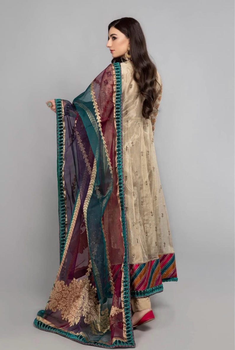 Pode incluir: Um vestido longo e fluido de cor bege com bordados dourados e uma bainha com uma faixa listrada colorida. O vestido &eacute; combinado com um dupatta transparente em tons de azul-turquesa, roxo e bord&ocirc;, com um design bordado em ouro e uma borda azul-turquesa.