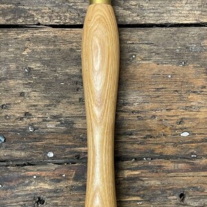 Op de afbeelding: Een houten handvat voor een snijgereedschap met een messing bus. Het handvat is gemaakt van lichtbruin hout met een nerfpatroon. De messing bus is glanzend en heeft een glad oppervlak.