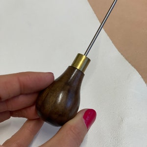 May include: A close-up of a leather stitching awl with a wooden handle and a brass ferrule. The awl is held in a hand with red nail polish.