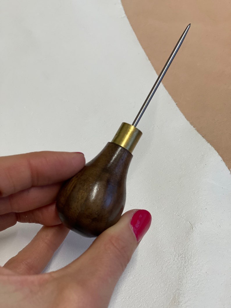 May include: A close-up of a leather stitching awl with a wooden handle and a brass ferrule. The awl is held in a hand with red nail polish.
