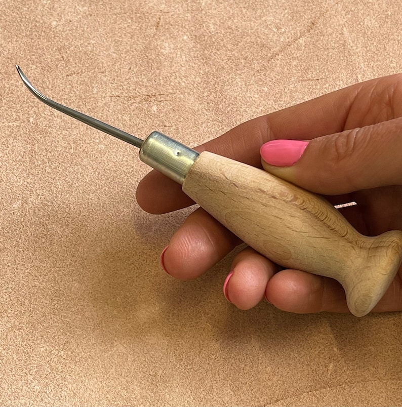 May include: A close-up of a leather sewing awl with a wooden handle. The awl has a curved needle and a metal ferrule.