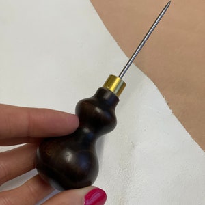 May include: A dark brown wooden handle with a brass ferrule and a long, thin, silver needle. The tool is used for leather crafting.