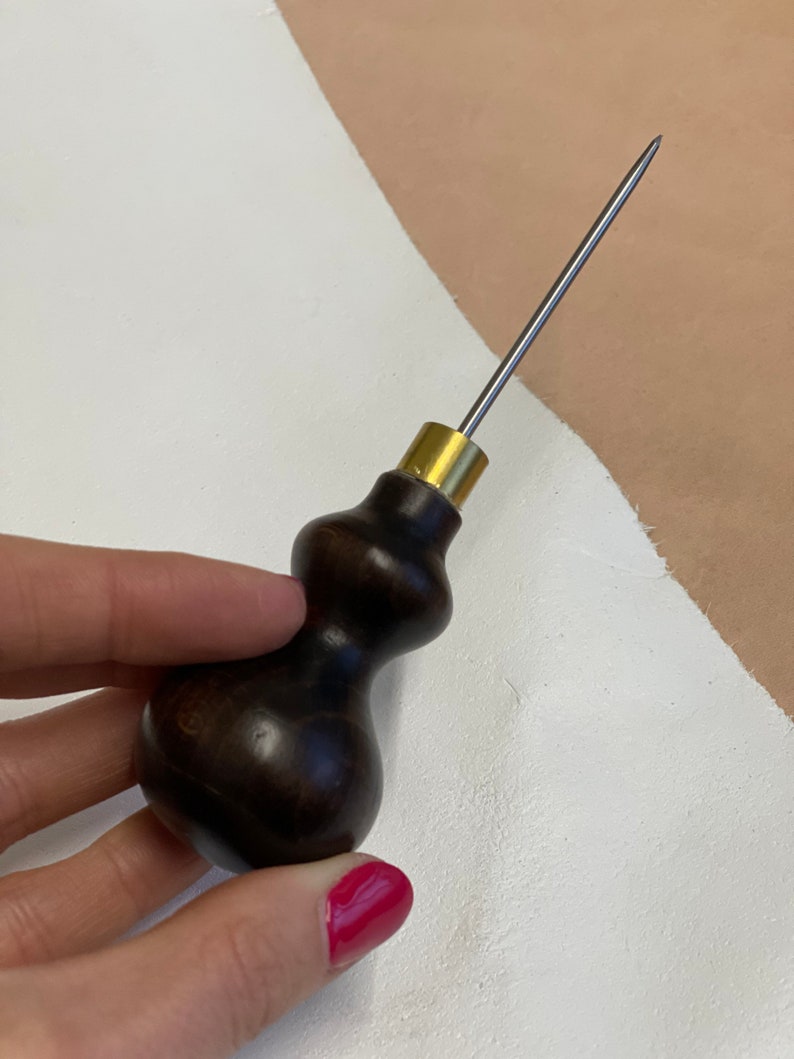 May include: A dark brown wooden handle with a brass ferrule and a long, thin, silver needle. The tool is used for leather crafting.