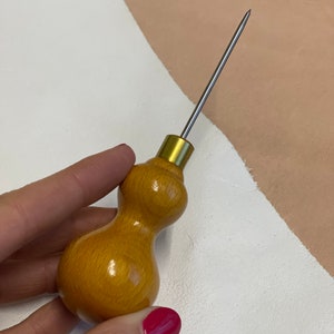 May include: A wooden leather stitching awl with a long, thin, metal needle. The handle is shaped like a gourd and is a light brown color.