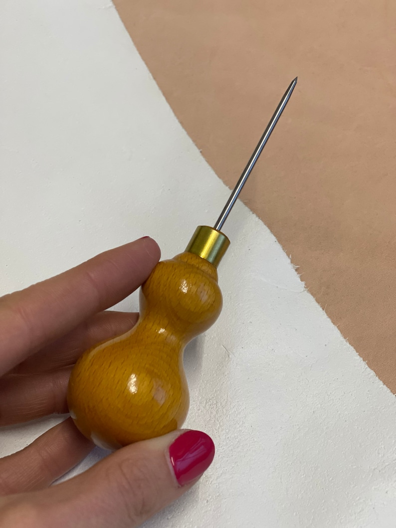 May include: A wooden leather stitching awl with a long, thin, metal needle. The handle is shaped like a gourd and is a light brown color.