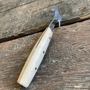 May include: A small, wooden-handled tool with a curved metal blade. The blade is sharp and pointed, and the handle is made of light wood with black dots.