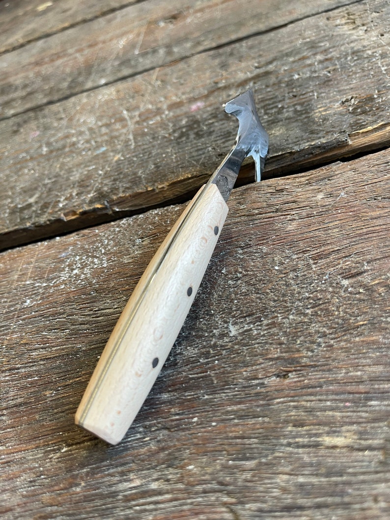 May include: A small, wooden-handled tool with a curved metal blade. The blade is sharp and pointed, and the handle is made of light wood with black dots.