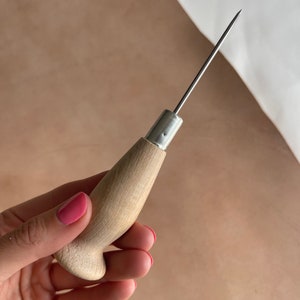 May include: A wooden leather stitching awl with a metal needle. The awl is held in a hand with pink nail polish.