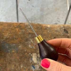 May include: A close-up of a leather awl with a dark wood handle and a brass ferrule. The awl is held in a hand with pink nail polish.