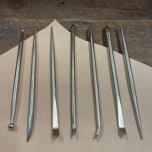 Puede incluir: Un conjunto de ocho herramientas de trabajo de cuero plateadas de diferentes formas y tamaños. Las herramientas están dispuestas sobre una superficie de color marrón claro.