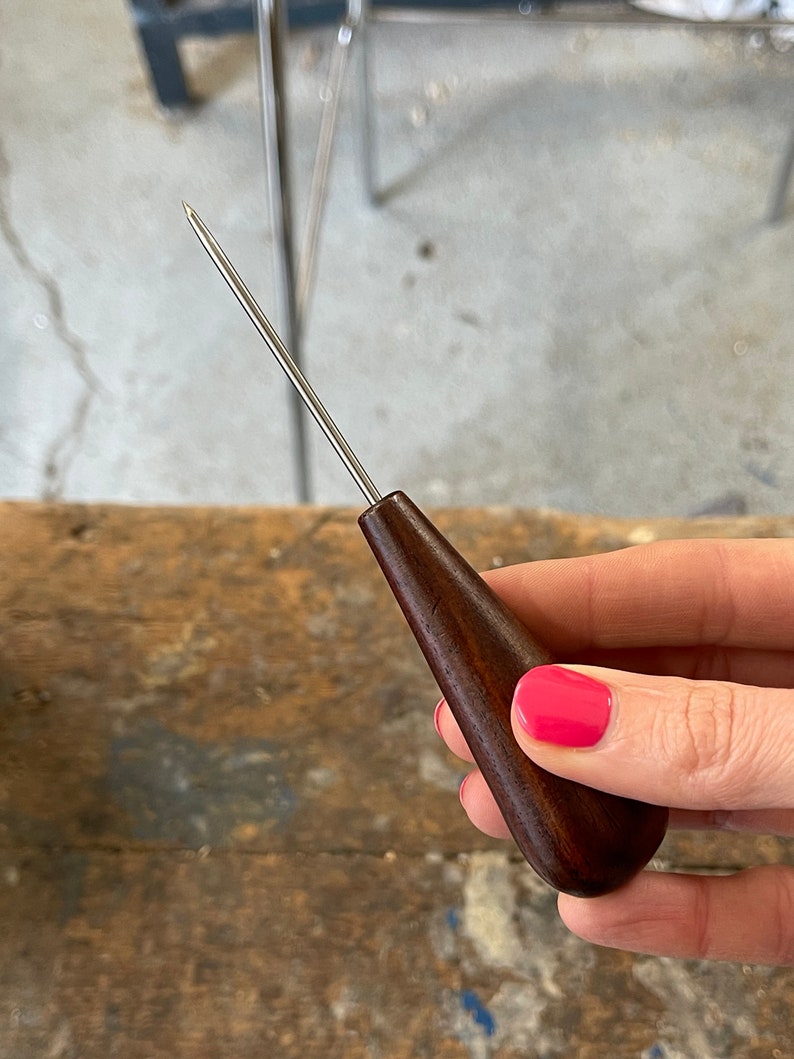 May include: A close-up of a wooden awl with a metal needle. The awl is held in a hand with pink nail polish.