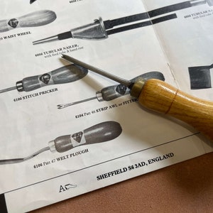 May include: A close-up of a set of leather working tools, including a stitch pricker, a strip awl, a welt plough, and a waist wheel. The tools are made of wood and metal and have a diamond-shaped logo on the handles. The tools are laid out on a white page with text that reads "6100 Stitch Pricker", "6104 Patt 46 Strip Awl or Fitter", "6106 Patt 47 Welt Plough", and "6006 Patt 28 Waist Wheel". The text also includes the company name and location: "Sheffield S6 3AD, England".