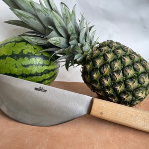 May include: A stainless steel curved knife with a light wood handle rests on a brown surface next to a green watermelon and a pineapple. The knife has the text "A.L. Columbus, Ohio" on the blade.