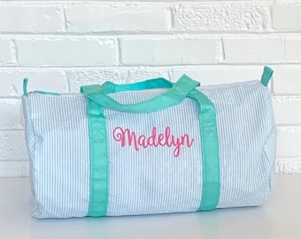 May include: A light blue and white striped duffel bag with a teal zipper and teal straps. The bag is personalized with the name "Madelyn" in pink.