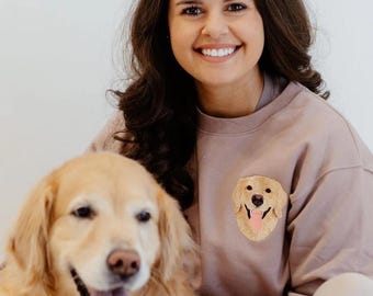 May include: A woman wearing a mauve sweatshirt with an embroidered golden retriever on the front, smiling and holding a golden retriever dog.