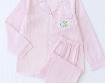 May include: Pink and white striped pajama set. The long-sleeved top has a button-down front, a chest pocket with green embroidered initials, and white trim. The matching pants are also striped.