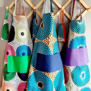 K&ouml;nnte beinhalten: Drei farbenfrohe Sch&uuml;rzen mit geometrischen Mustern h&auml;ngen an einem Holzgestell. Die Sch&uuml;rzen haben unterschiedlich farbige Taschen und sind aus einem Stoff mit kreisf&ouml;rmigem Muster.