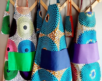 Könnte beinhalten: Drei farbenfrohe Schürzen mit geometrischen Mustern hängen an einem Holzgestell. Die Schürzen haben unterschiedlich farbige Taschen und sind aus einem Stoff mit kreisförmigem Muster.