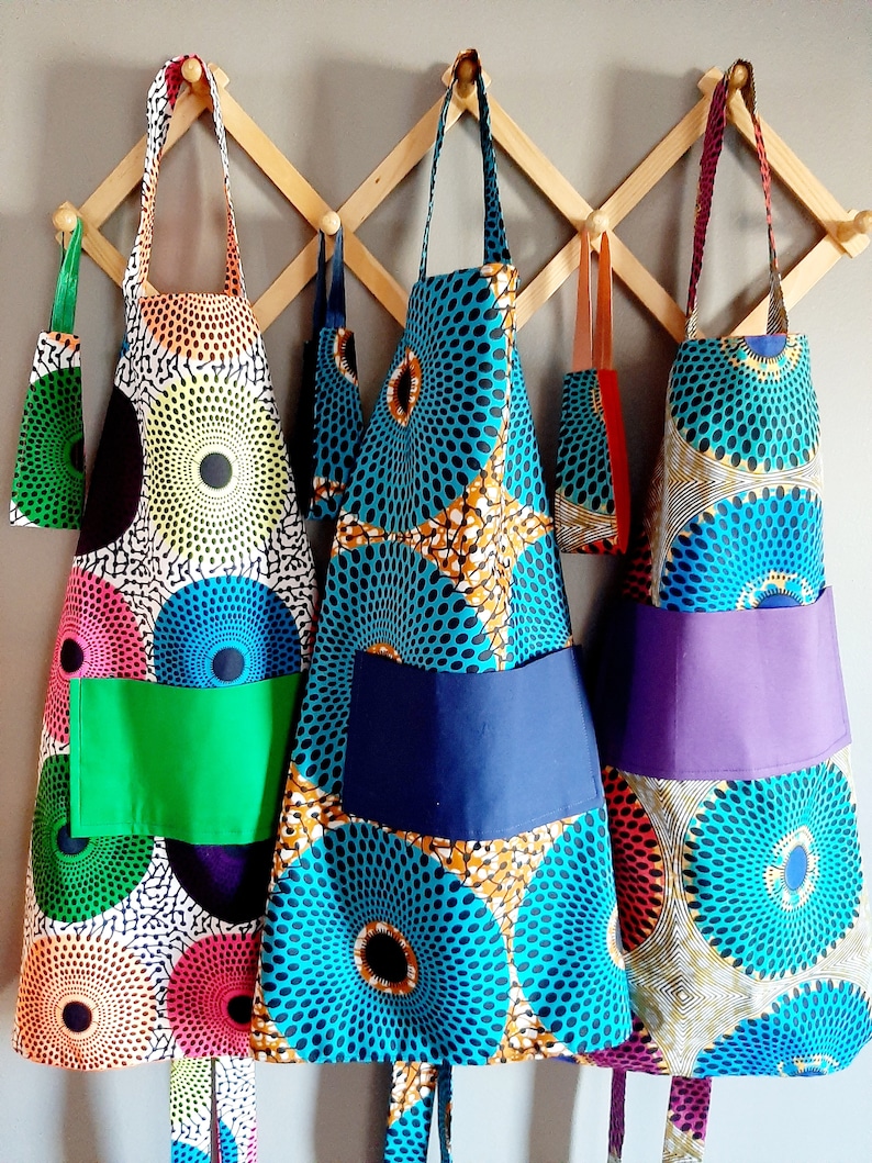 K&ouml;nnte beinhalten: Drei farbenfrohe Sch&uuml;rzen mit geometrischen Mustern h&auml;ngen an einem Holzgestell. Die Sch&uuml;rzen haben unterschiedlich farbige Taschen und sind aus einem Stoff mit kreisf&ouml;rmigem Muster.