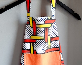 Könnte beinhalten: Eine farbenfrohe Schürze mit einem geometrischen Muster in Rot, Gelb und Blau auf weißem Hintergrund. Sie hat eine große orangefarbene Tasche auf der Vorderseite. Die Schürze hängt an einem schwarzen Kleiderbügel mit den Buchstaben LMM.