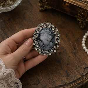 May include: An oval brooch featuring a grey cameo of a profile portrait of a person with curly hair and a rose, surrounded by a decorative silver-toned metal frame encrusted with small clear rhinestones arranged in a circular pattern.