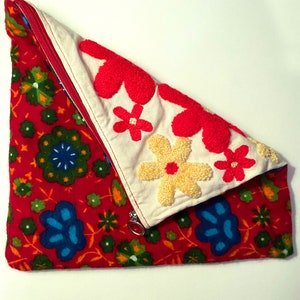 May include: A red fabric pouch with a zipper closure and a white fabric flap with embroidered flowers in red, yellow, and white. The pouch is decorated with a floral pattern in red, green, and blue.