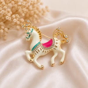May include: A gold-tone metal brooch shaped like a carousel horse, featuring white enamel, a bright pink saddle, and teal accents. The horse is decorated with clear rhinestones along the mane, saddle, and tail, with a polished gold finish.