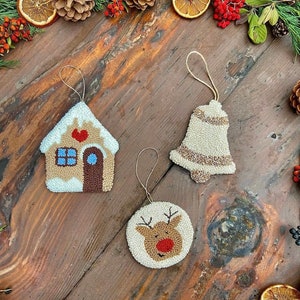 Op de afbeelding: Drie handgemaakte kerstversieringen: een gingerbreadhuis, een bel en een rendier. Elke versiering is gemaakt van wit garen en heeft kleurrijke details.