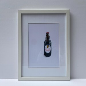 May include: A framed print featuring a Guinness stout bottle. The bottle is dark brown with a red cap and a white label with the Guinness logo. The print is in a white frame, set against a light gray background.