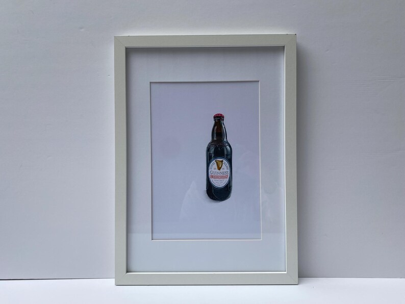 May include: A framed print featuring a Guinness stout bottle. The bottle is dark brown with a red cap and a white label with the Guinness logo. The print is in a white frame, set against a light gray background.
