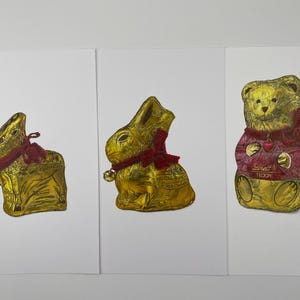 Puede incluir: Tres figuras de chocolate envueltas en papel de aluminio: un conejo, un conejo y un oso de peluche. Los conejos tienen bufandas y campanas rojas. El oso de peluche lleva un su&eacute;ter rojo con la palabra "Teddy".