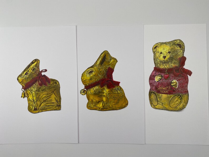 Puede incluir: Tres figuras de chocolate envueltas en papel de aluminio: un conejo, un conejo y un oso de peluche. Los conejos tienen bufandas y campanas rojas. El oso de peluche lleva un su&eacute;ter rojo con la palabra "Teddy".