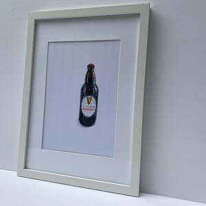 May include: A framed print featuring a Guinness stout bottle. The bottle is dark with a red cap and a white label with the Guinness logo. The print is in a white frame, set against a white background.