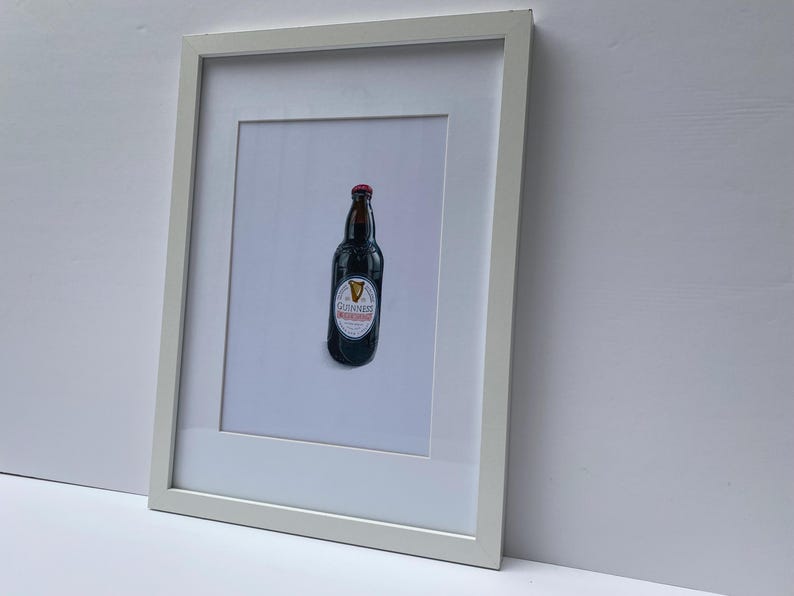 May include: A framed print featuring a Guinness stout bottle. The bottle is dark with a red cap and a white label with the Guinness logo. The print is in a white frame, set against a white background.