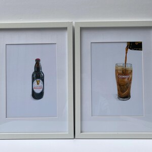 May include: Two framed prints featuring Guinness beer. One print shows a bottle of Guinness with a red cap and a white label. The other print depicts beer being poured into a glass, with the Guinness logo visible.