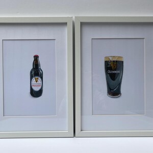 May include: Two framed prints featuring Guinness. One print shows a bottle of Guinness with a red cap and a white label. The other print shows a pint of Guinness with the Guinness logo.