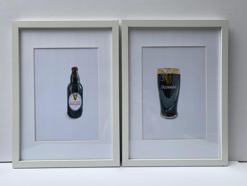 May include: Two framed prints featuring Guinness. One print shows a bottle of Guinness with a red cap and a white label. The other print shows a pint of Guinness with the Guinness logo.