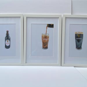 May include: Three framed prints featuring Guinness beer. One shows a bottle, another pouring beer, and the last a full pint. The prints are in white frames, with the Guinness logo visible on each.