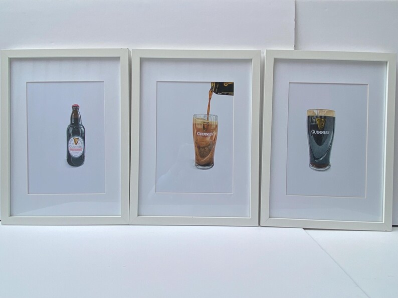 May include: Three framed prints featuring Guinness beer. One shows a bottle, another pouring beer, and the last a full pint. The prints are in white frames, with the Guinness logo visible on each.