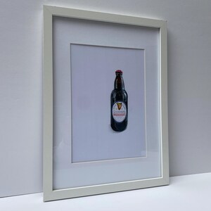 May include: Framed print featuring a dark brown Guinness beer bottle with a red cap and a white and red label. The framed artwork is displayed against a white background, encased in a white frame.